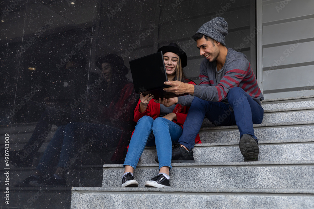 Two young Latin American brothers sitting on the stairs with laptop and cell phone while sharing some work time watching videos and messaging. Technology concept. Two young Latin American brothers sitting on the stairs with laptop and cell phone while sharing some work time watching videos and messaging. Technology concept.