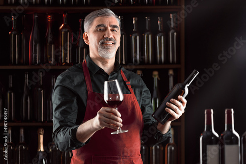 Winemaker examinating wine products among shelves of wine bottles. Stylish middle-aged man with a grey beard