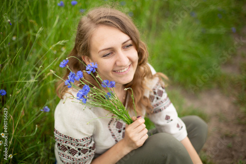 Girl wearing national Ukrainian clothes embroidered shirt is sitting in the beautiful rye field. Woman is holding blue field flowers cornflowers. Blue wildflowers and blond wavy haired girl 