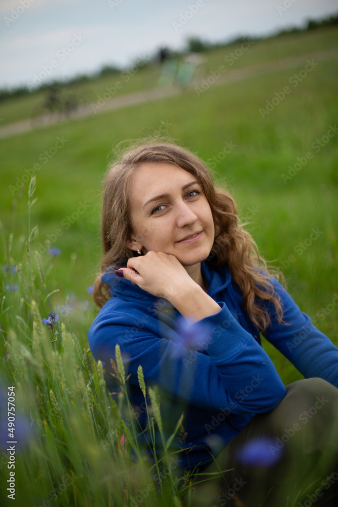 Satisfied blond girl is sitting on green grass near blue wildflowers. Women is wearing dark blue hoodie near cornflowers on the green background.
Satisfied blond girl is sitting on green grass near blue wildflowers. Women is wearing dark blue hoodie near cornflowers on the green background.