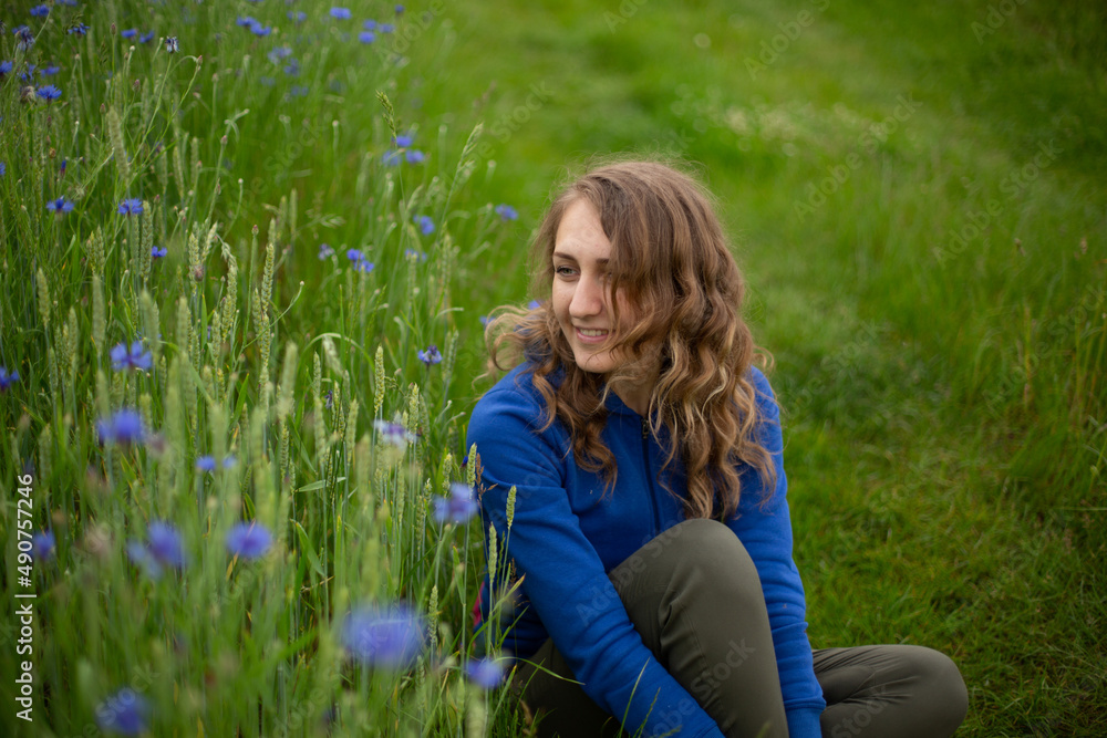Fototapeta premium Satisfied blond girl is sitting on green grass near blue wildflowers. Women is wearing dark blue hoodie near cornflowers on the green background. 