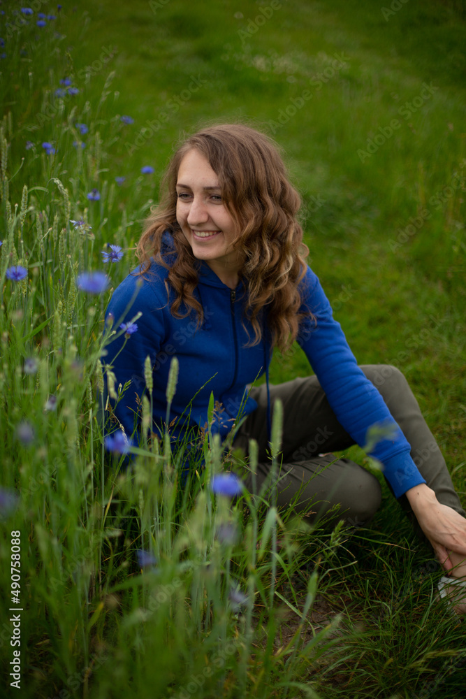 Satisfied blond girl is sitting on green grass near blue wildflowers. Women is wearing dark blue hoodie near cornflowers on the green background.
Satisfied blond girl is sitting on green grass near blue wildflowers. Women is wearing dark blue hoodie near cornflowers on the green background.