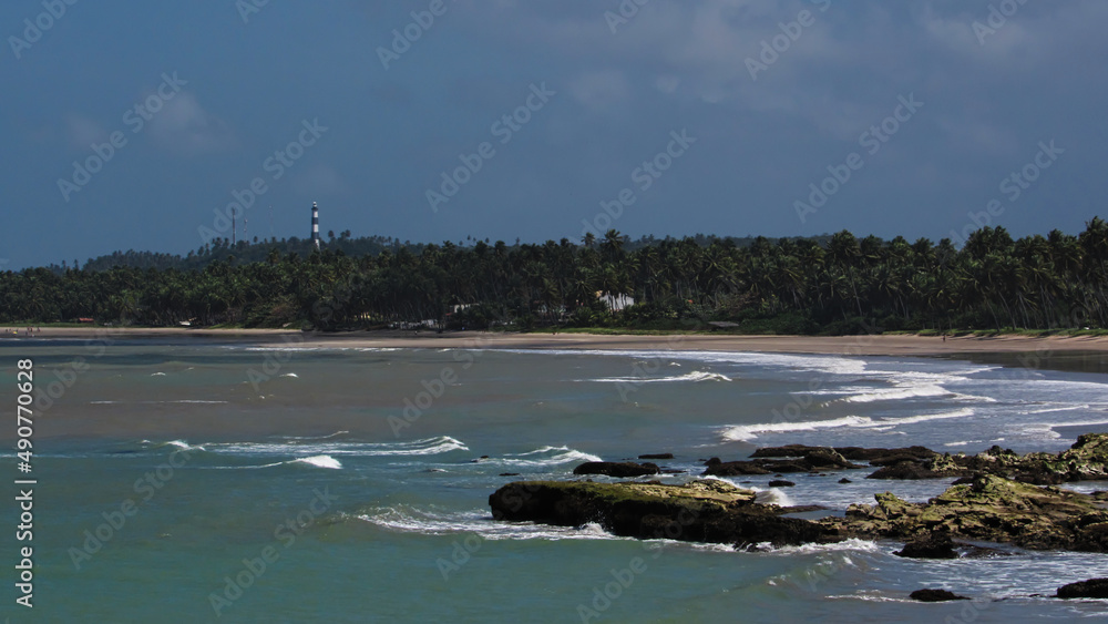 visible rocks in the low tide and to the bottom crashing waves in the sand of the beach, coconut forest and lighthouse tower illuminated by the sun, under a misty sky, Costa dos Corais, AL, Brazil visible rocks in the low tide and to the bottom crashing waves in the sand of the beach, coconut forest and lighthouse tower illuminated by the sun, under a misty sky, Costa dos Corais, AL, Brazil