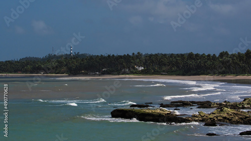 visible rocks in the low tide and to the bottom crashing waves in the sand of the beach, coconut forest and lighthouse tower illuminated by the sun, under a misty sky, Costa dos Corais, AL, Brazil photo