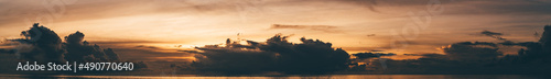 A panoramic view of a stunning dramatic sunset over the ocean, evening on the Maldives, tropical warm sunlight around dark silhouettes of clouds, a water lever at the bottom