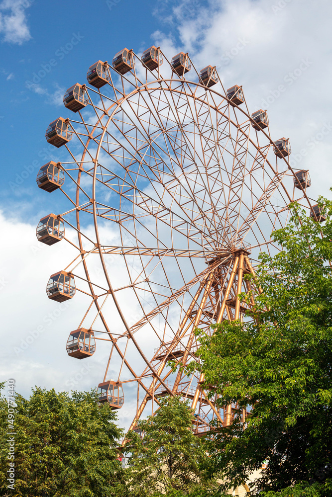 A big observation wheel in an amusement park against a blue sky with clouds. A vertical image. A big observation wheel in an amusement park against a blue sky with clouds. A vertical image.