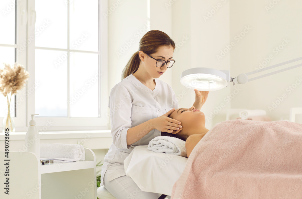 Female client who wants to clean her face came to beautician's office for cosmetic procedures. Beautician examines client under lamp while she lies on couch during facial treatment at beauty clinic. Female client who wants to clean her face came to beautician's office for cosmetic procedures. Beautician examines client under lamp while she lies on couch during facial treatment at beauty clinic.
