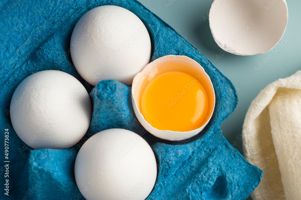 Close-up of chicken fresh eggs in eco-packaging on a blue background. Broken egg with yolk in the shell. Farm natural products. Top view. - obrazy, fototapety, plakaty 