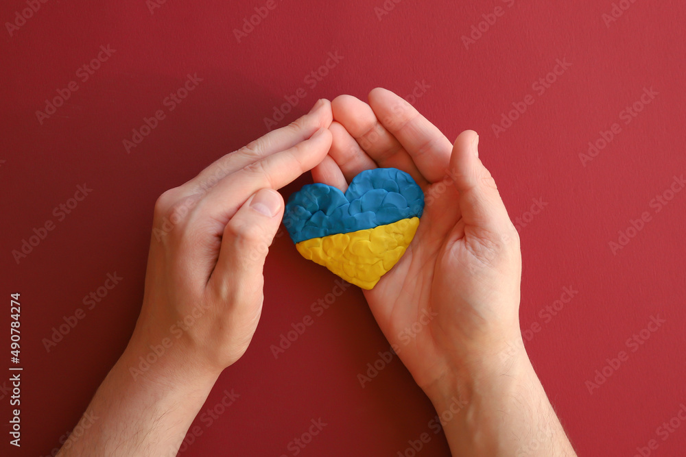 Ukrainian Ukraine flag, no war. Heart shape of blue and yellow plasticine modeling clay in male hands on red maroon texture background. Plasticine finger textured background. Top view, closeup. Ukrainian Ukraine flag, no war. Heart shape of blue and yellow plasticine modeling clay in male hands on red maroon texture background. Plasticine finger textured background. Top view, closeup.
