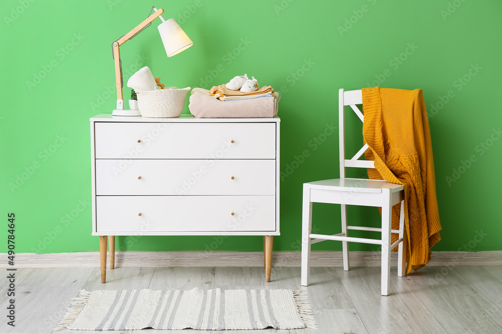 Fototapeta premium Chest of drawers with baby clothes and chair with knitted sweater near color wall in room interior