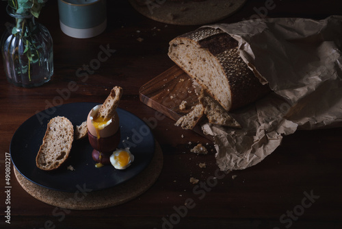 Breakfast table scene. Chiaroscuro style food photo. Cracked and open boiled egg with dippy bread set against a dark background with copy space available
 photo