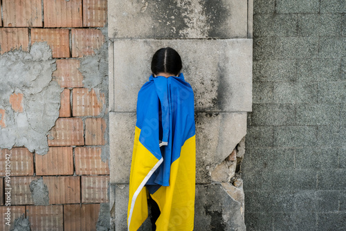 Little girl with Ukrainian flag in front of a wall destroyed from bombs. The little girl waves the national flag while praying for peace