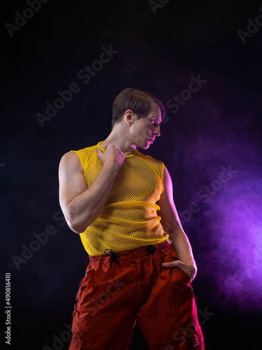 a young muscular man in a bright outfit poses in a pretentious pose, style and grotesque. Attractive guy with pumped-up arms in a mesh T-shirt and red pants, photo in smoke