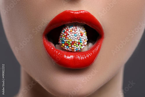 Young woman with red lips makeup eating candy on grey background, closeup