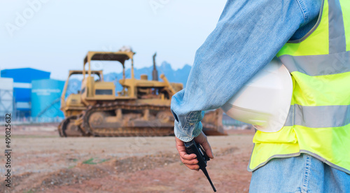 An Asian male engineer or worker wearing a vest holding a radio transmitter and a helmet, a middle-aged foreman controls the excavator.