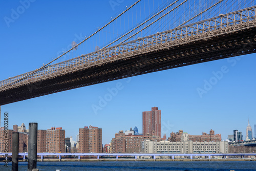 Looking up at the span section of Brooklyn Bridge on a sunny day with blue sky. High-rise residential buildings behind the suspension bridge deck © MichaelVi