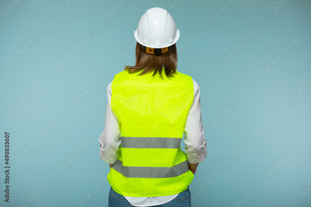 Professional engineer. A woman in a protective helmet and a bright vest stands with her back to the camera, against a blue background. Copy paste. Professional engineer. A woman in a protective helmet and a bright vest stands with her back to the camera, against a blue background. Copy paste.