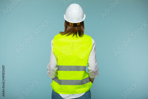 Professional engineer. A woman in a protective helmet and a bright vest stands with her back to the camera, against a blue background. Copy paste.
