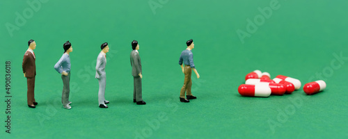 There are pills on the green surface  to which there is a line of miniature figures of people. Banner.