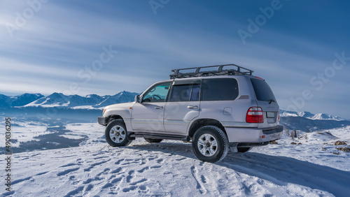 The SUV car stands on a snowy plateau. Footprints and shadows in the snow. Ahead and below you can see the river, the mountain range against the blue sky. Altai