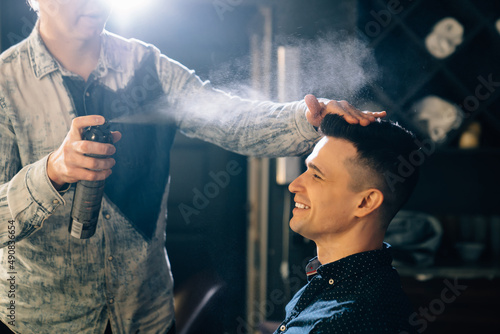 the hairdresser styling a brunette man and spraying hairspray in a barbershop photo