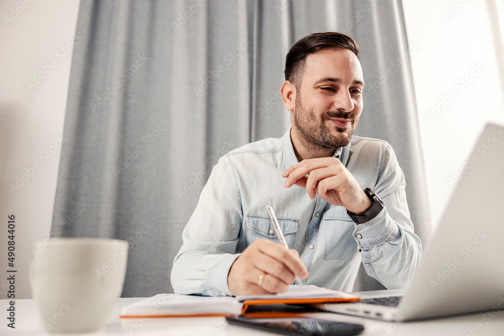 Naklejka premium A freelancer looking at the laptop, writing down, working at home office.