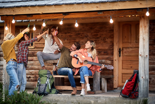Fototapeta Naklejka Na Ścianę i Meble -  Group of young friends toasting and playing guitar in front of wooden cottage on the terrace.  Summertime garden celebration and fun. Friends, togetherness, fun, joy, celebration concept.