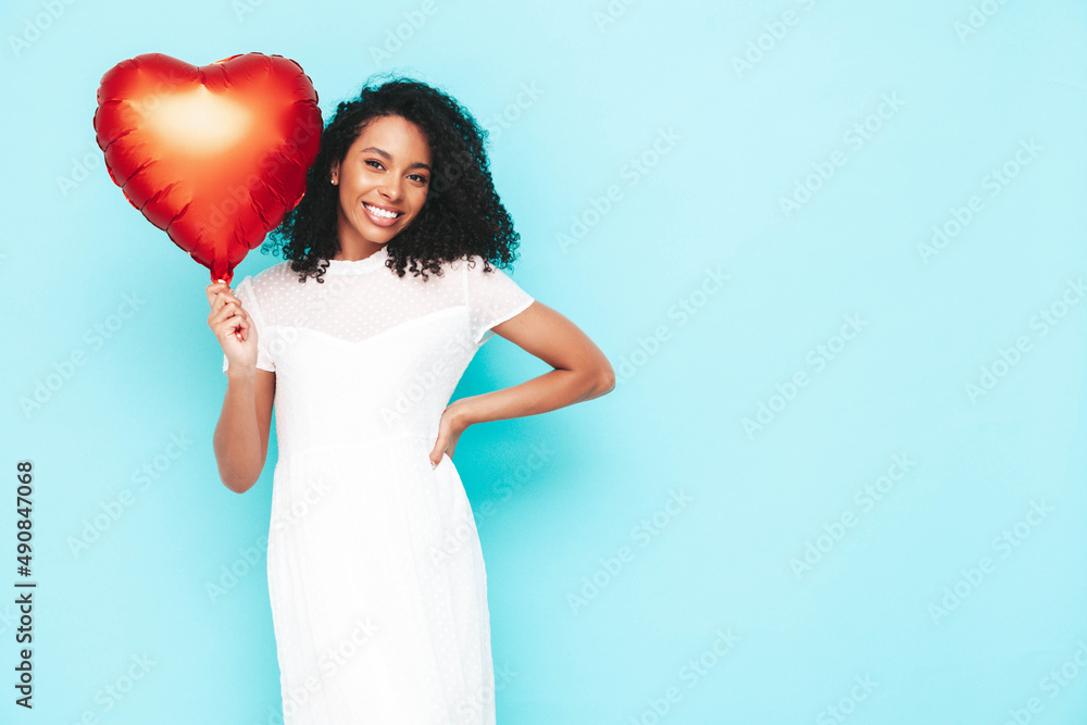 Beautiful black woman with afro curls hairstyle. Smiling model dressed in white summer dress. Sexy carefree female posing near blue wall in studio. Tanned and cheerful. Holding heart air balloon Beautiful black woman with afro curls hairstyle. Smiling model dressed in white summer dress. Sexy carefree female posing near blue wall in studio. Tanned and cheerful. Holding heart air balloon