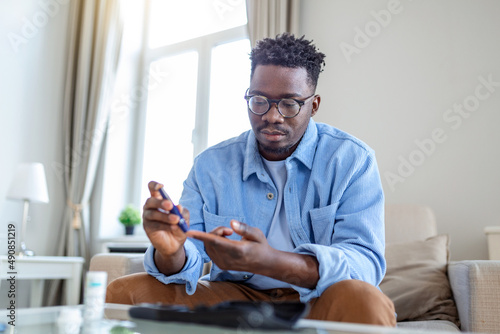 African man is sitting at the sofa and taking blood from his finger due to diabetes. The daily life of a man of African-American ethnicity person with a chronic illness who is using glucose tester. photo