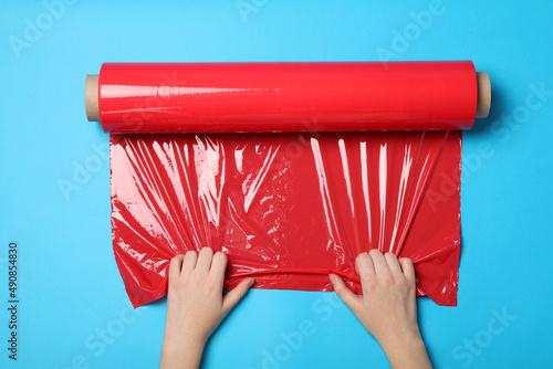 Woman unrolling red plastic stretch wrap on light blue background, top view photo