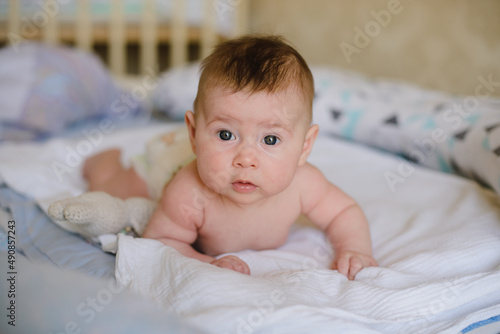 The baby is lying on his stomach on the bed on a white sheet. Looks at the camera and smiles.