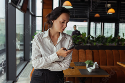 Confident female restaurant manager in formal outfit browsing smartphone while working in hall and standing near table 