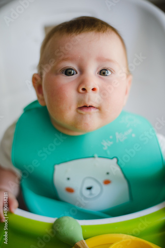 A baby in a bib is sitting on a high chair in front of the table, smiling, eating mashed potatoes with a spoon. Looks at the camera