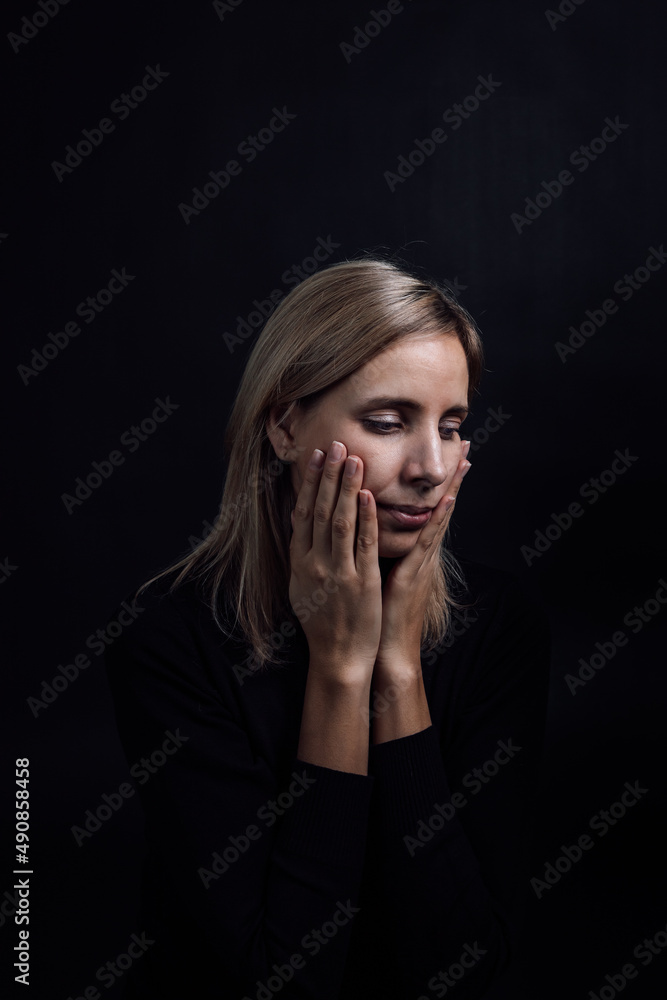 Pretty depressed woman with hands on cheeks in black dress on black background. Victim of physical and psychological abuse. Gaslighting. Relative aggression. Pretty depressed woman with hands on cheeks in black dress on black background. Victim of physical and psychological abuse. Gaslighting. Relative aggression.