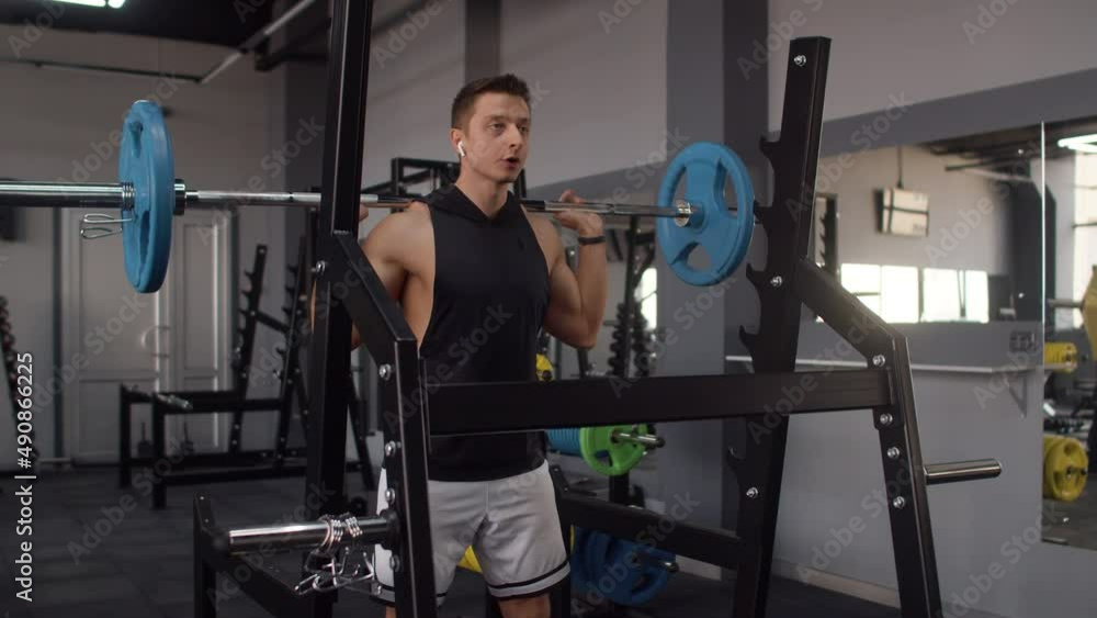 Man of athletic build in sportswear and headphones trains at gym doing squats with barbell, front view. Man of athletic build trains in gym with barbell Man of athletic build in sportswear and headphones trains at gym doing squats with barbell, front view. Man of athletic build trains in gym with barbell