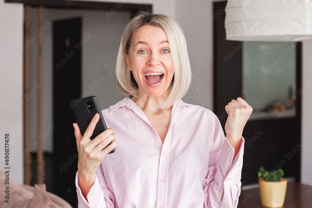 Attractive middle-aged 40s woman looking at her smart phone and smiling while standing at home Attractive middle-aged 40s woman looking at her smart phone and smiling while standing at home