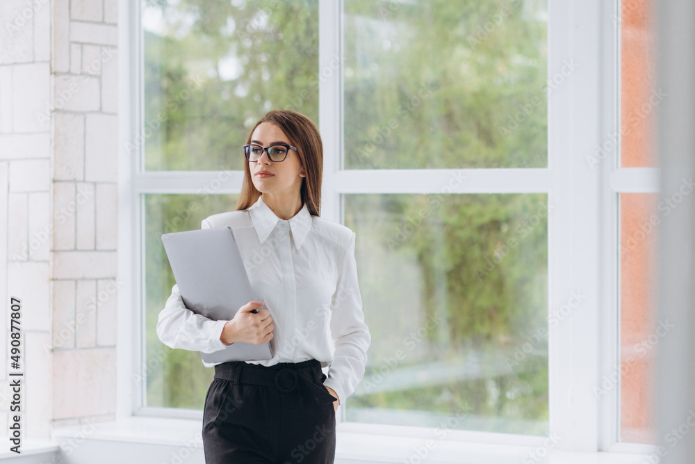 young successful girl standing in the office and holding a laptop. The girl is dressed in a white shirt and black pants young successful girl standing in the office and holding a laptop. The girl is dressed in a white shirt and black pants