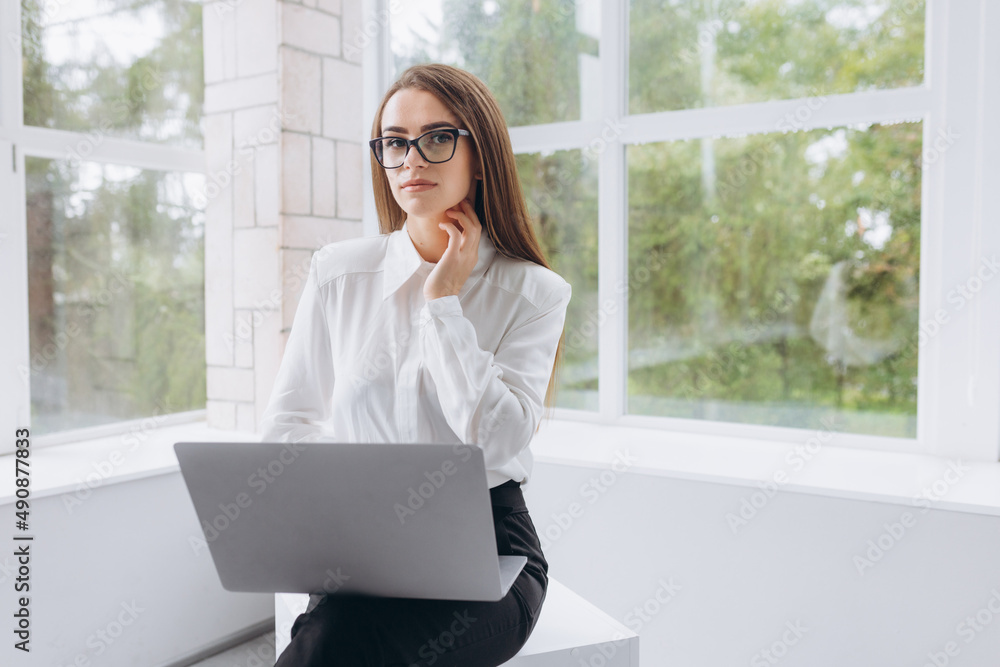 Portrait of a young beautiful business woman in the office. The girl is wearing a white shirt with glasses. The girl works at the laptop Portrait of a young beautiful business woman in the office. The girl is wearing a white shirt with glasses. The girl works at the laptop