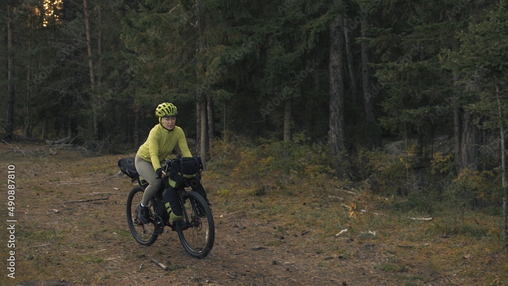 Naklejka premium The woman travel on mixed terrain cycle touring with bike bikepacking outdoor. The traveler journey with bicycle bags. Stylish bikepacking, bike, sportswear in green black colors. Magic forest park.