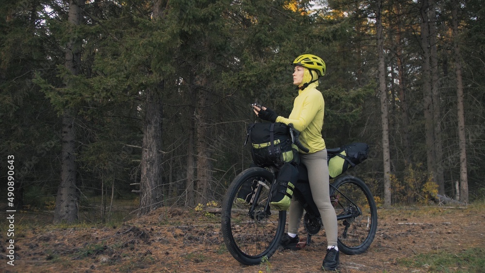 The woman travel on mixed terrain cycle touring with bike bikepacking outdoor. The traveler journey with bicycle bags. Sportswear in green black colors. Magic forest park. Make a selfie smartphone. The woman travel on mixed terrain cycle touring with bike bikepacking outdoor. The traveler journey with bicycle bags. Sportswear in green black colors. Magic forest park. Make a selfie smartphone.