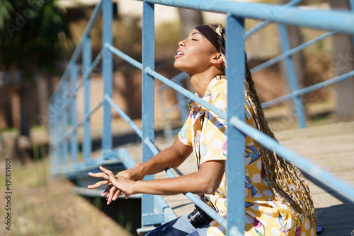 Black girl with braids sitting on a bridge enjoying the sun on her face with her eyes closed.