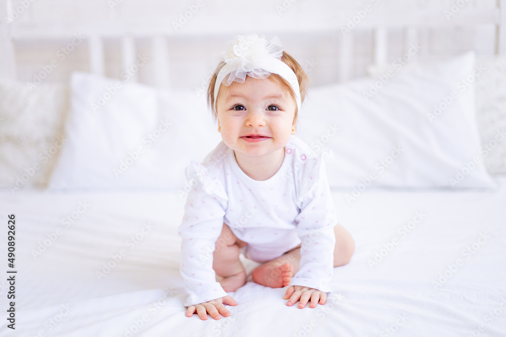 cute baby girl sitting on the bed in white clothes and with a bow on her head, funny little baby on a cotton bed at home and smiling, baby products concept cute baby girl sitting on the bed in white clothes and with a bow on her head, funny little baby on a cotton bed at home and smiling, baby products concept