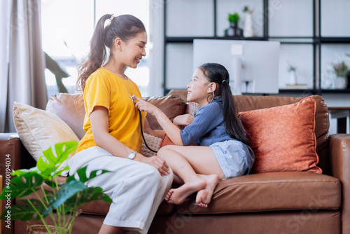 asian daughter play doctor checking up her mother with care and happiness in living room on sofa couch at home,female daughter playroll doctor nurse use stethoscope listen her mom heart pulse with fun