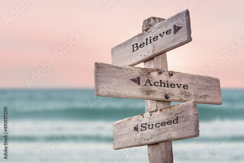 believe achieve suceed text quote written on wooden signpost by the sea. Positive pink turqoise pastel theme. photo