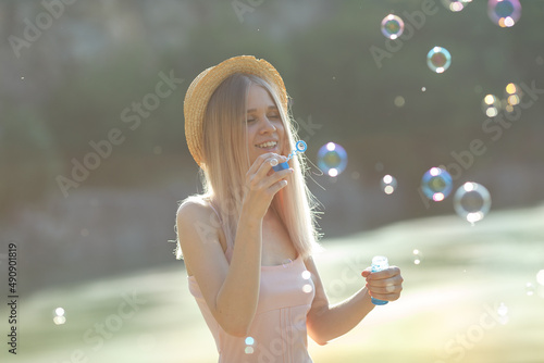 young attractive blonde girl in a straw hat and dress blows soap bubbles on the shore of the lake. Summer mood