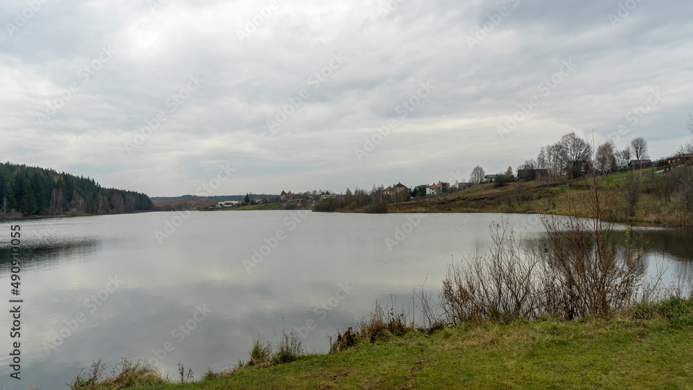 On the shore of a forest lake. Grey clouds and the trees reflected symmetrically in the water. Lake beach panorama.