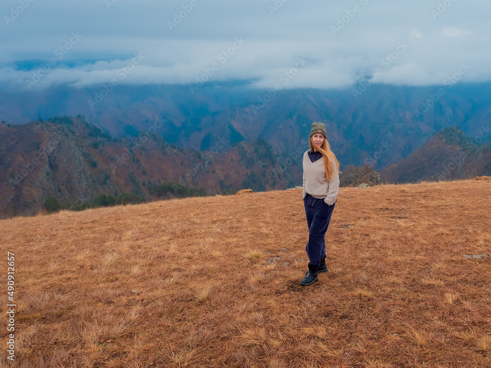 A woman walks on dry grass in a field located in the Caucasian highlands with clouds covering the peaks.