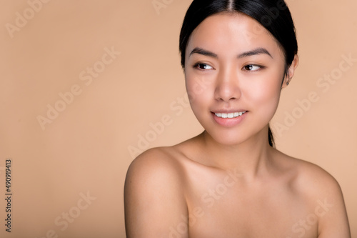 Beauty and health concept. A beautiful Asian girl with perfect, well-groomed facial skin, with nude shoulders, stands on an isolated beige background, looks away, smiles. Copy space, mock-up concept