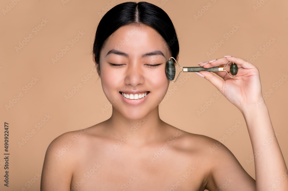 Fototapeta premium Asian beautiful girl holding a jade massage roller for facial skin in her hand, makes anti-aging measures, wrinkle prevention, standing on an isolated beige background, smiling with eyes closed