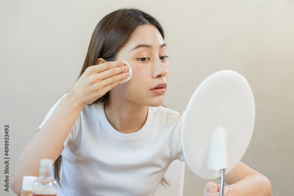 Happy beauty, beautiful asian young woman, girl looking in to mirror, holding cotton pad, applying facial wipe on her face, removing makeup before shower in bathroom, skin care on white background. Happy beauty, beautiful asian young woman, girl looking in to mirror, holding cotton pad, applying facial wipe on her face, removing makeup before shower in bathroom, skin care on white background.